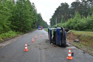 Samochód który brał udział w kolizji drogowej. Uszkodzony pojazd znajduje się na jezdni, jest przewrócony na bok. W tle wóz strażacki.
