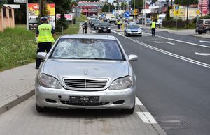 Po lewej stronie znajduje się samochód koloru szarego. Pojazd ma pękniętą szybę. Obok samochodu stoi policjant