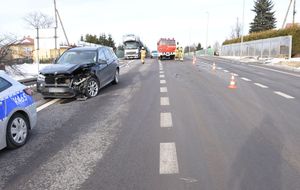 Miejsce zderzenia pojazdów. Po lewej stronie drogi znajduje się uszkodzony pojazd. W tle służby ratownicze