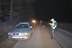 Policjant na drodze podczas wykonywania czynności służbowych na miejscu wypadku drogowego. Po lewej stronie na poboczu znajduje się samochód.