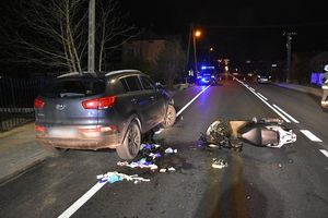 Zdarzenie drogowe. Po lewej stronie znajduje się samochód osobowy. Po prawej stronie na jezdni leży motocykl uszkodzony w wyniku zdarzenia.