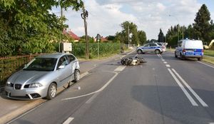 Miejsce zdarzenia drogowego. Ulica na której na poboczu znajduje się samochód osobowy, na środku jezdni leży motocykl, a w tle radiowóz blokujący drogę.