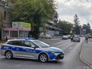 Miejsce wypadku drogowego. Na drodze przed przejściem dla pieszych znajduje się radiowóz. Za przejściem samochód osobowy obok którego stoi policjant.