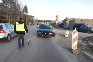 Policjant - technik kryminalistyki wykonuje czynności na miejscu wypadku drogowego. W tle uszkodzony w wyniku zdarzenia pojazd ososbowy