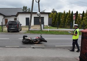 Miejsce wypadku drogowego. Na ulicy znajduje się przewrócony motocykl. Przed nim stoi policjant