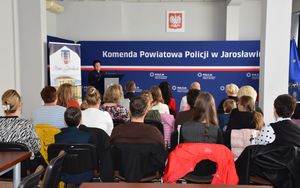 Aula na której osoby siedzą na krzesłach. W tle policjantka przy mównicy