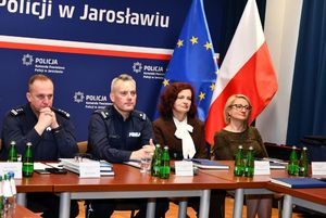 Policjanci i dwie kobiety podczas odprawy służbowej. W tle flagi Polski i Unii Europejskiej.