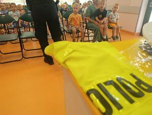 Policyjna kamizelka odblaskowa. W tle policjantka podczas prelekcji z dziećmi